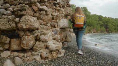 Genç bir kadın antik bir binanın taş duvarı boyunca ilerliyor ve dalgalar neredeyse duvarlarına değiyor..