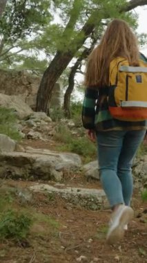 Dikey video. Genç ve yalnız bir kadın gezgin, deniz kıyısındaki bir kayanın kenarındaki ormanın içinden geçerek antik bir şehrin kalıntıları olan taşların üzerinden geçiyor..