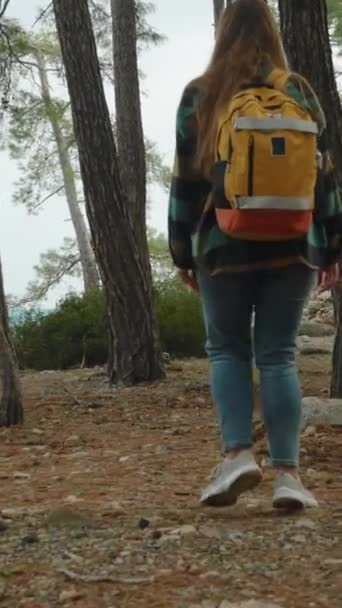バーティカルビデオ 若い女性の背後からバックパック付きの平らなシャツを見ると 海と古代遺跡で森の中を一人でハイキングして旅します — ストック動画
