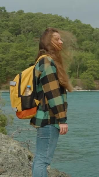垂直录像 一个年轻女子乱蓬蓬的长发在风中飘扬 孤身一人背着背包旅行海边凉爽的天气 悬崖峭壁上的茂密森林 — 图库视频影像
