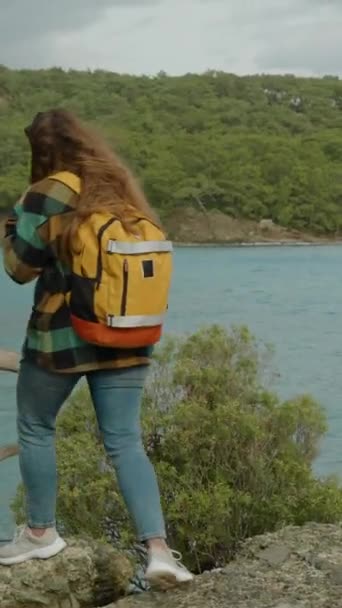 バーティカルビデオ バックパックを持つ若い女性観光客は 古くて乾燥した木のトランクに沿って崖から海に降りてきます ブルーウォーターの湾の眺め — ストック動画
