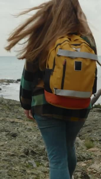 垂直录像 一个背着背包的女游客爬上岩石 享受大海和凉爽的风 寂寞和忧郁阴郁的天气 — 图库视频影像