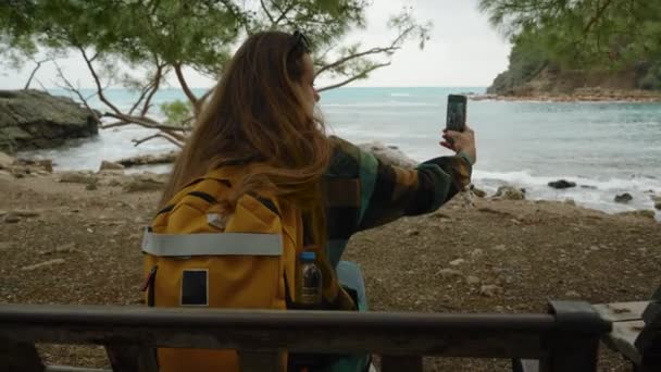 若い女性 旅行ブロガーは ソーシャルメディアのセルフィー写真を撮るか 保管庫として撮ります 海を望む松の木の下のベンチでリラックス — ストック動画