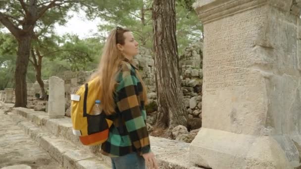 古代リシアの街 ファシリス バックパックに触れた若い女性観光客は 古代ギリシャ語の碑文で石柱を調べます — ストック動画