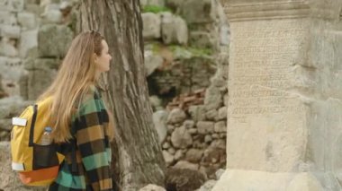 Dişi gezgin, antik kentteki harabelerin arka planına kazınmış yazıtlarla antik bir Yunan taş sütununu inceliyor..