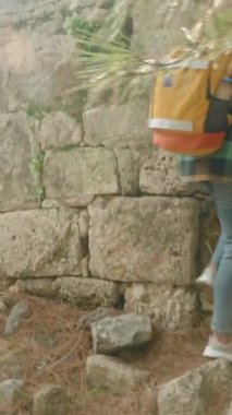Dikey video. Sırt çantalı bir kadın gezgin çam ormanındaki antik bir şehrin taşlarının ve kalıntılarının arasına tırmanıyor. Antik Yunan evleri.