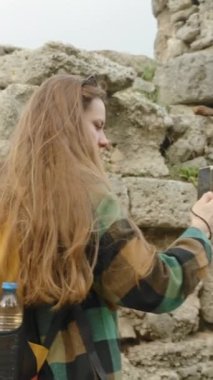 Dikey video. Genç bir kadın milenyum gezgini veya seyahat blogcusu sosyal medya veya hatıra olarak ormandaki antik bir Yunan amfi tiyatrosunun fotoğraflarını çekiyor.