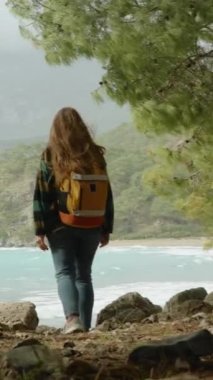 Dikey video. Kamera, çam ağaçlarında yürürken sırt çantalı bir kadını takip ediyor, taşlara basıyor ve turkuaz suyla birlikte koyda beliriyor. Rüzgarlı bir hava. Yalnızlık ve kasvetli bir ruh hali. Yavaş çekim.