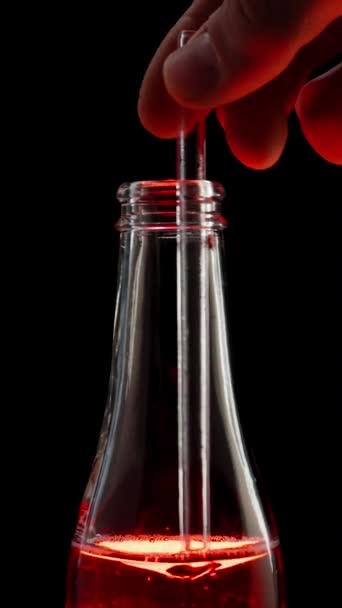 垂直录像 玻璃瓶中的红色苏打水用透明的吸管搅拌 在里面形成涡旋 在黑色背景下 — 图库视频影像