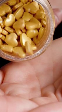 Dikey video. Altın gibi küçük kalp şeklinde şekerler, bir kadın bunları kavanozdan eline döker. Pasta süslemesi için şeker parçacıkları.