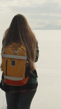 Dikey video. Deniz kıyısındaki bir uçurumun kenarında sırt çantalı genç ve yalnız bir kadın yolcunun manzarası. Ellerini katladı..