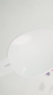 Dikey video. Çok renkli kalpler yukarıdan bir kaşığa düşer. Beyaz arkaplan, pasta şefi için şeker serpiştirme. Yavaş çekim.