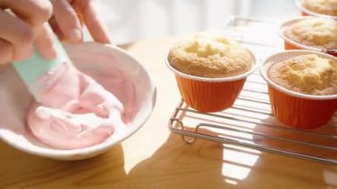 Parlak güneş ışığı altında, ahşap bir masanın üzerinde keklerin hazırlanması, bir kadın pembe kremayı karıştırıyor. Dekorasyon için şekerler yakınlarda..