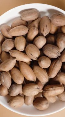 Dikey video. Fındıklı fındık kabuklu beyaz tabağın yukarıdan aşağı görünümünü açık kahverengi bir arkaplanda döndürmek.