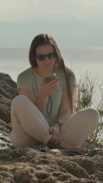 バーティカルビデオ 若い女性は海で岩の上に座って電話を見る — ストック動画