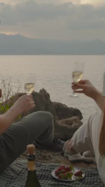 Dikey video. Genç Çiftin Romantik Buluşması Battaniyeye Otururlar, Şampanya Kadehleri Çıngırdatırlar ve Denizde Gün Batımına Hayran kalırlar.