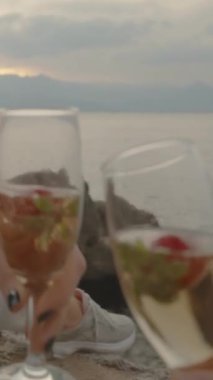Dikey video. Birinci Şahıs Manzarası, Bayanla Şampanyayı Çakışan Bardaklar, Deniz kenarında Battaniyede Oturmak ve Sunset 'i izlemek. Bardakta Çilek.