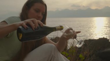 Genç kadın bir bardağa şampanya dolduruyor denizin arka planına ve dağlarda gün batımına karşı. Yavaş çekim.