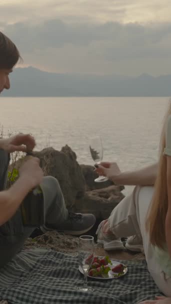 垂直录像 男人为女人倒香槟 日落日期在海边的岩石上 覆盆子上的草莓 — 图库视频影像