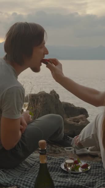 バーティカルビデオ 恋人のペア 海でクリフにブランケットに座る 女性は男のストロベリーをフィードし 彼らはシャンパンを飲む サンセットを提唱 — ストック動画