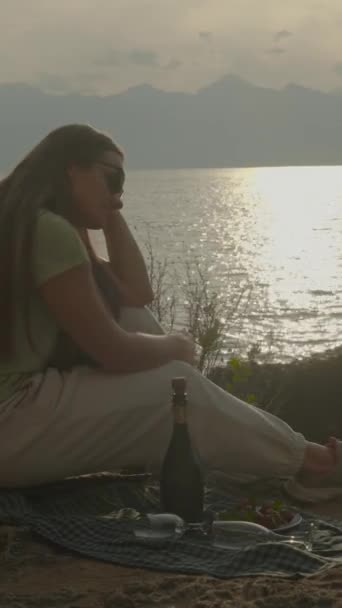 垂直录像 日落时 年轻的女人悲伤地坐在海边 等待着男人的约会 盖着眼镜的香槟 — 图库视频影像