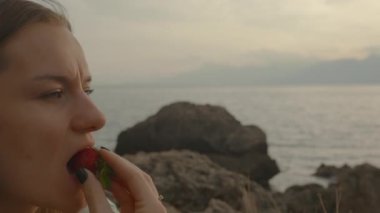 Uzun saçlı kadın gün batımında deniz kenarındaki bir kayanın üzerinde oturmuş çilek yiyor..
