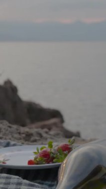 Dikey video. Randevudan sonra boş şampanya şişesi ve çilekli bardaklar. Denizde gün batımının arkaplanına karşı.