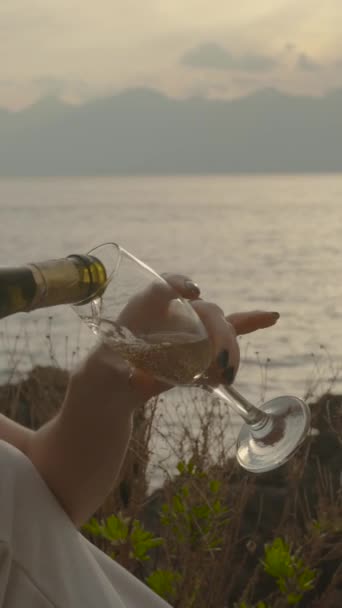 バーティカルビデオ 若い女性は 山の海と日没の背景に対して シャンパンをガラスに注いでいます スローモーション — ストック動画