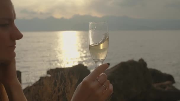 女人在杯子里旋起香槟酒 看着夕阳西下的阳光反射在海面上 — 图库视频影像