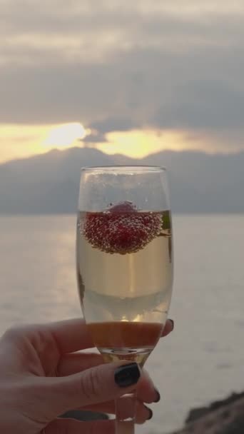 バーティカルビデオ 女性の手は 海の山の背後にある設定の太陽の雨の背景に対して 泡の中のイチゴとシャンパングラスを保持しています — ストック動画