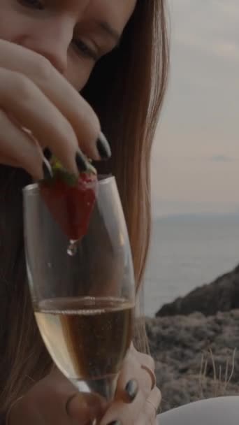 垂直录像 年轻女人从香槟酒杯中取出草莓 吃了它 然后在海上的日落前把它洗干净 — 图库视频影像