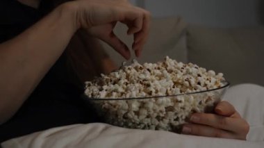 Genç bir kadın, karanlık bir odada televizyonun karşısında otururken patlamış mısır yiyor..