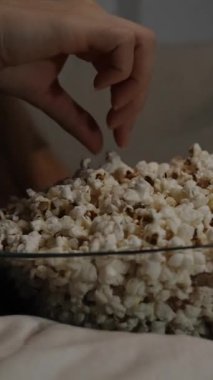 Dikey video. Genç bir kadın, karanlık bir odada televizyonun karşısında otururken patlamış mısır yiyor..