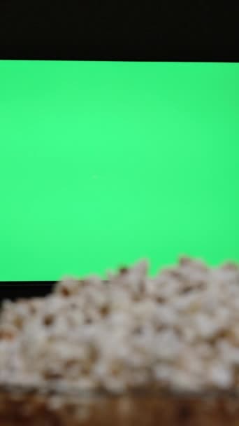 垂直录像 一只手从电视机前的碗里摸到爆米花 用一个空的绿色屏幕的模型 — 图库视频影像