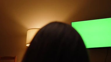Yeşil Ekranlı Uzun Saçlı Bir Kadının Başının Arkasından TV İzle. Lambadan gelen ısıtma odası ışıkları..