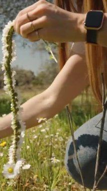 Dikey video. Uzun saçlı genç bir bayan turist parkta beyaz çiçeklerden oluşan bir çelenk dokuyor ve uzaktaki dağlarda da dağlar var. Güneş ışınları, dairesel hareket.