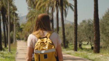 Uzun saçlı ve sırt çantalı bir kadın palmiye ağaçlarıyla uzun bir yol boyunca dağlara doğru yürüyordu. Arka görünüm.