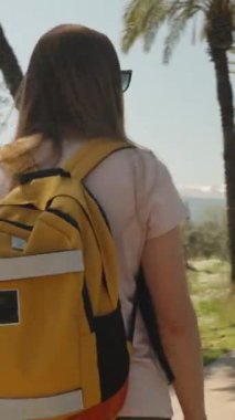 Dikey video. Kamera sırt çantalı kadını takip ediyor ve dairesel hareket ediyor. Yalnız bir yolculuk, uzun palmiyelerin yanında yol..