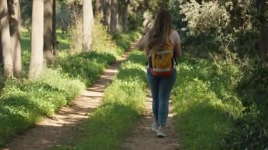 Kamera, güneşli orman yolunda sırt çantalı bir kadını takip ediyor..