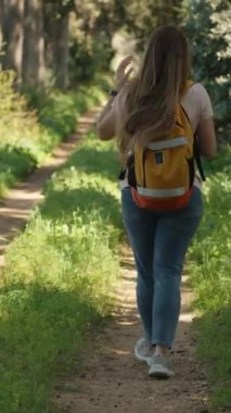 Dikey video. Uzun saçlı ve kafasında papatya çelengi olan genç bir kadın sırt çantasıyla ormanda yürüyor..