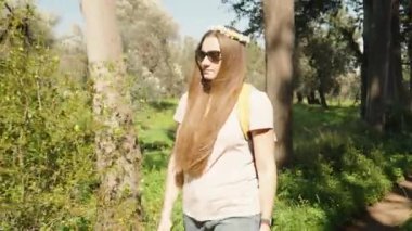 Uzun saçlı ve kafasında papatya çelengi olan genç bir kadın sırt çantasıyla ormanda yürüyor..