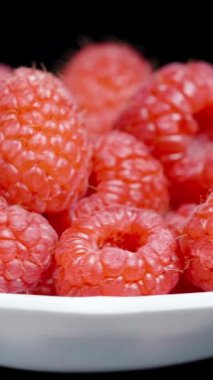 Dikey video. Küçük beyaz bir tabakta ahududu, siyah bir arka planda döngü içinde dönüyor..