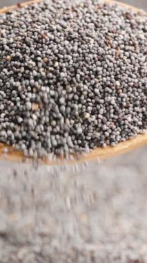 Dikey video. Arka planda haşhaş tohumları ve ön plandaki tahta kaşıktan dökülen bir avuç haşhaş tohumu..