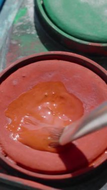Dikey video. Bir suluboya palet, kırmızı boyaya batırılmış bir fırça. Yakın çekim, pürüzsüz.