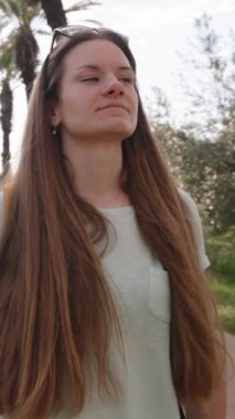 Dikey video. Sırt çantalı uzun saçlı bir kadın, arkasında güneş ışınları olan palmiyeler arasında yol boyunca seyahat eder. Yavaş çekim, dairesel hareket.