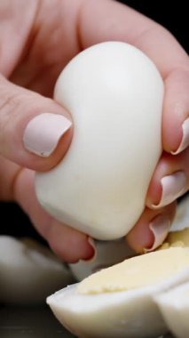 Dikey video. Bir kadının eli haşlanmış bir yumurtayı sıkar ve her yöne doğru uçan küçük parçalara ayırır. Ağır çekim. Siyah bir arkaplana karşı.