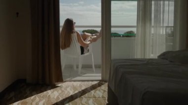 Beyaz yataklı bir otel odası ve balkonda güneşlenen bir kadın ve deniz manzarasına hayran kalarak şampanyanın tadını çıkarıyorlar..