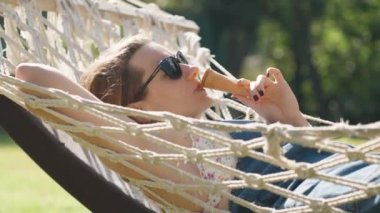 A young woman basks in the sunlight while swaying in a hammock and eating ice cream.
