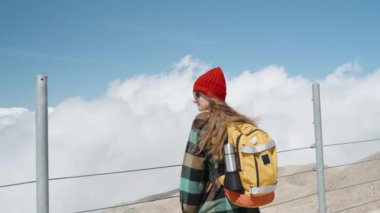 A young and solitary female traveler in a red hat and with a backpack stands on the cool summit of the mountain, admiring the views of the clouds.