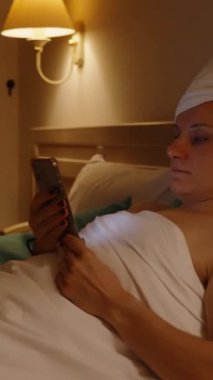 Vertical video. A Woman Rests on the Bed After a Shower in the Hotel. She Looks at Her Phone as the Camera Moves in a Circle, Capturing the Interior of the Room.
